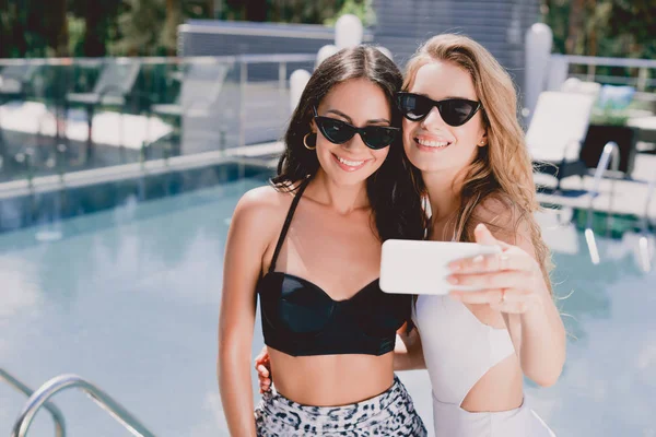 Sorridente loira e morena amigos em óculos de sol e maiôs tomando selfie perto da piscina — Fotografia de Stock