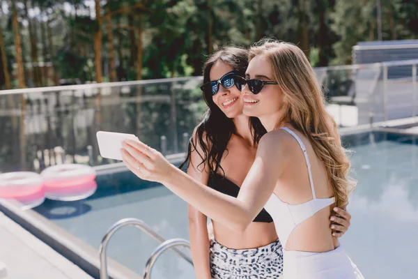 Heureuses femmes blondes et brunes en lunettes de soleil et maillots de bain prenant selfie près de la piscine — Photo de stock