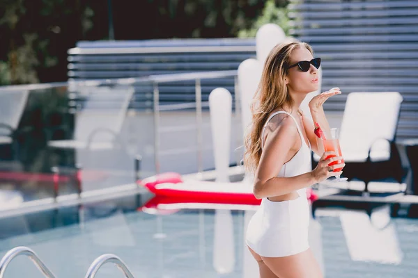 Sexy blonde woman in white swimsuit with cocktail blowing air kiss near swimming pool — Stock Photo