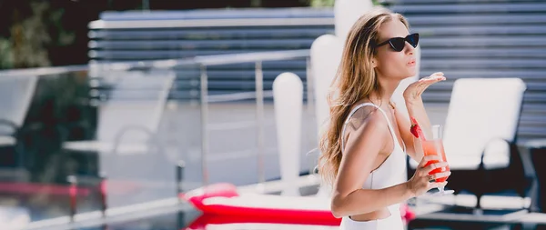 Sexy blonde woman in white swimsuit with cocktail blowing air kiss near swimming pool, panoramic shot — Stock Photo