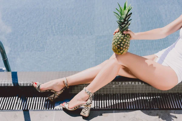 Cropped view of sexy woman sitting near swimming pool in white swimsuit and sandals with pineapple — Stock Photo