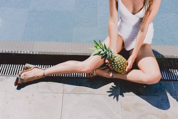 Cropped view of sexy woman sitting near swimming pool in white swimsuit with pineapple — Stock Photo