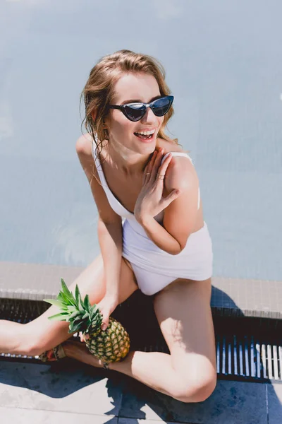Happy blonde woman in sunglasses and swimsuit sitting with pineapple near swimming pool — Stock Photo
