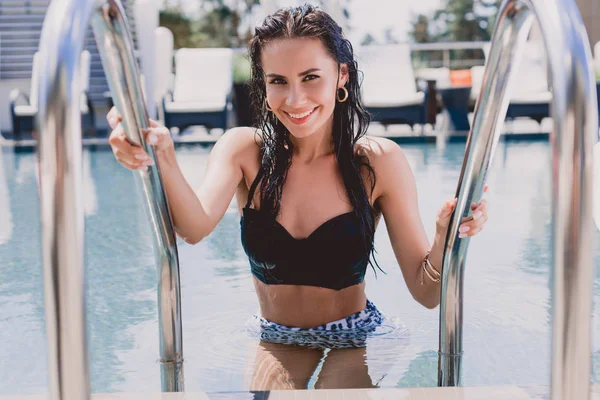 Selective focus of happy wet brunette woman with hands on metal handrails getting out from water in swimming pool — Stock Photo