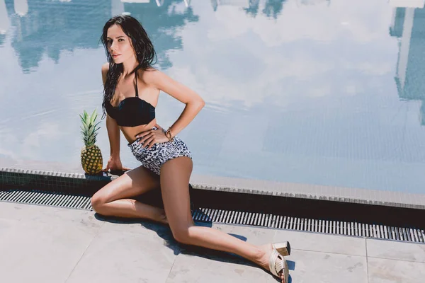 Sexy brünette Frau in Badeanzug und Sandalen posiert in der Nähe von Schwimmbad und Ananas — Stockfoto