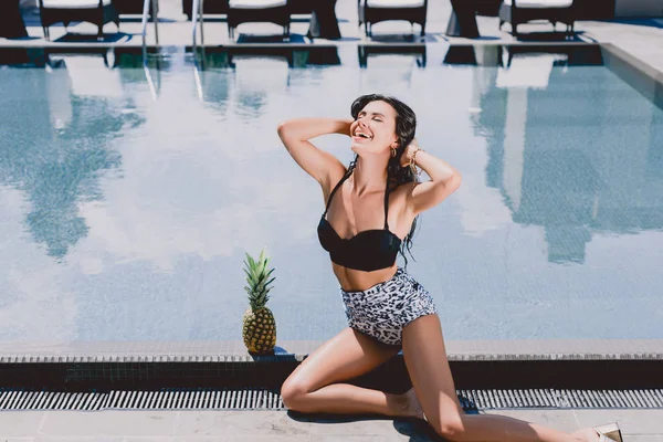 Счастливая сексуальная брюнетка в купальнике сидит с закрытыми глазами возле бассейна и ананаса — стоковое фото