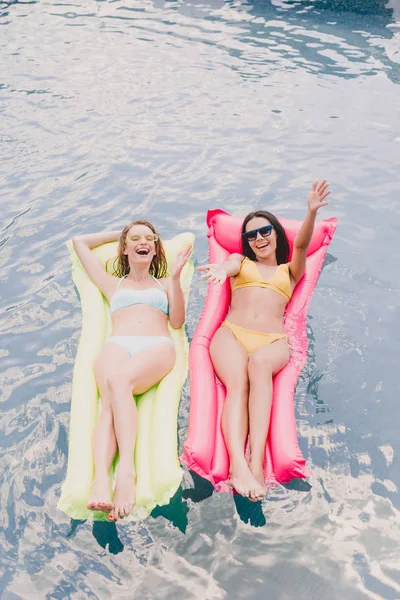 Vista aérea de morena e loira mulheres felizes e excitadas deitadas em flutuadores de piscina com as mãos estendidas na piscina — Fotografia de Stock