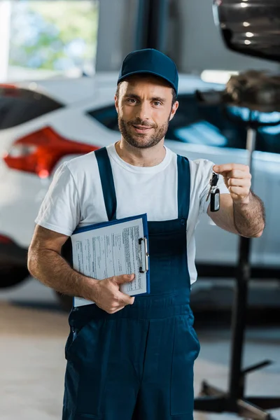 Felice meccanico auto guardando la fotocamera e tenendo appunti e chiave vicino auto — Foto stock