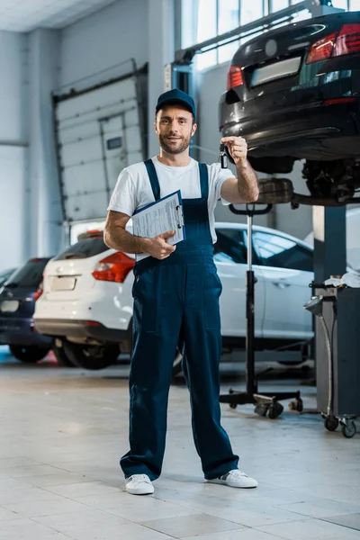 Happy car mechanic looking at camera and holding clipboard and key near cars — Stock Photo