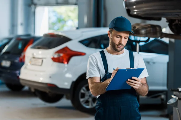 Bearded car mechanic writing while holding clipboard near cars — Stock Photo