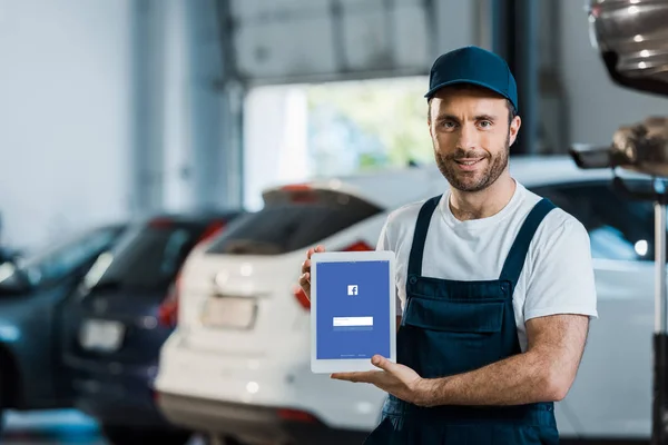 KYIV, UKRAINE - 7 JUIN 2019 : joyeux mécanicien de voiture tenant tablette numérique avec application facebook à l'écran dans le service de voiture — Photo de stock