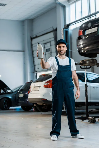 Heureux mécanicien de voiture dans le bouchon tenant bouteille avec de l'huile de voiture tout en se tenant en service de voiture — Photo de stock