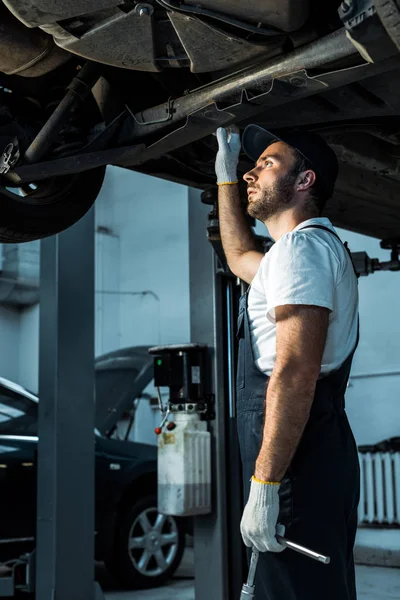 Bearded car mechanic in cap standing near automobile in car service — Stock Photo