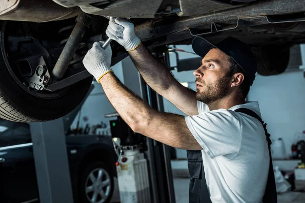 Bearded auto mechanic in cap repairing automobile in car service — Stock Photo