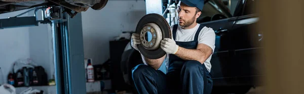 Panoramic shot of handsome car mechanic looking at metallic car brake — Stock Photo