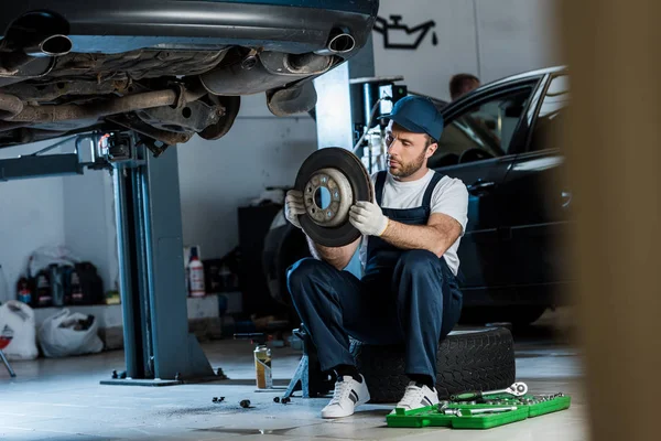 Selective focus of handsome car mechanic looking at metallic car brake — Stock Photo