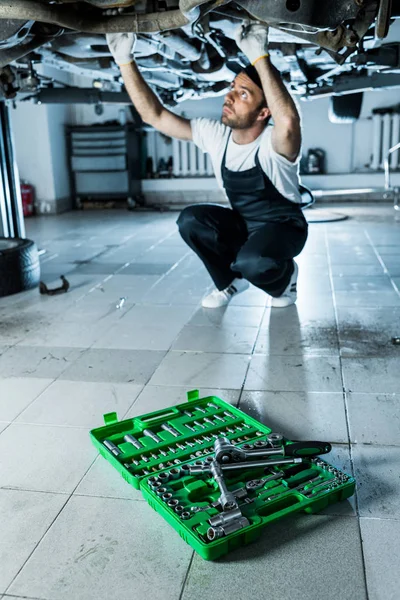 Selective focus of toolbox with tools near handsome car mechanic repairing automobile — Stock Photo