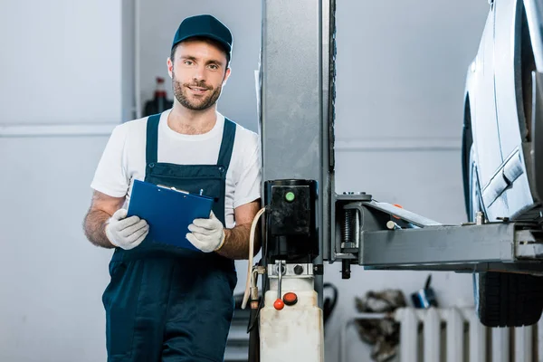 Cheerful auto mechanic in cap holding clipboard in car service — Stock Photo