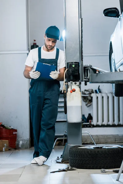 Bearded auto mechanic in cap looking at clipboard in car service — Stock Photo