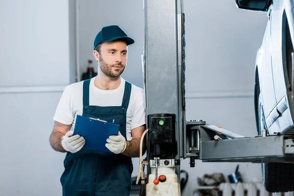 Bearded auto mechanic in cap looking at car and holding clipboard in car service — Stock Photo