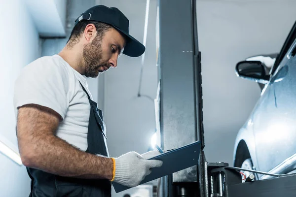 Vue à angle bas du mécanicien automatique barbu dans le chapeau regardant le presse-papiers dans le service de voiture — Photo de stock