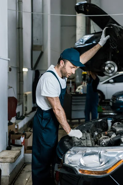 Красивый бородатый автомеханик в перчатках глядя на двигатель автомобиля — стоковое фото