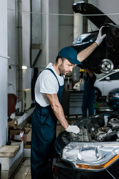 Beau barbu mécanicien automobile dans des gants regardant moteur de voiture — Photo de stock