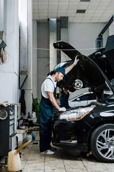 Handsome bearded auto mechanic in gloves and cap looking at car engine — Stock Photo