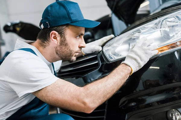 Handsome bearded auto mechanic in cap looking at tail light in black car — Stock Photo
