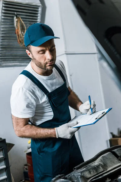 Selective focus of handsome repairman in cap holding pen and clipboard  near car — Stock Photo