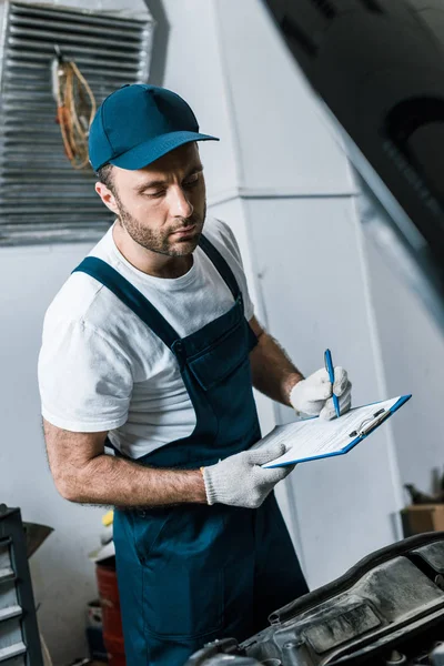 Selektiver Fokus des gutaussehenden Reparateurs in Mütze mit Stift und Klemmbrett in der Nähe des Autos — Stockfoto