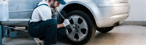 Panoramic shot of handsome bearded car mechanic in uniform changing car tire in car service — Stock Photo