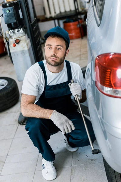 Selective focus of bearded car mechanic in uniform holding metallic wrench near car — Stock Photo