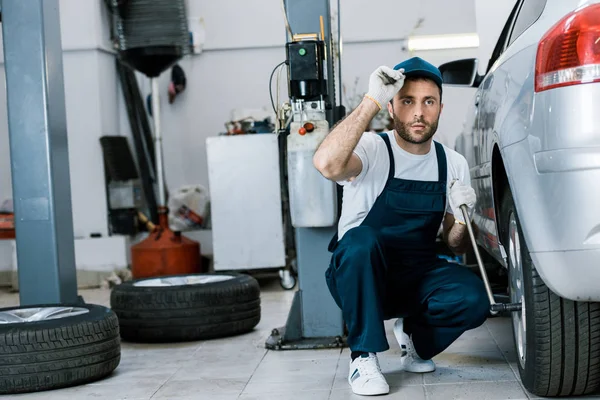 Schöner Automechaniker in Uniform hält Metallschlüssel neben Auto — Stockfoto