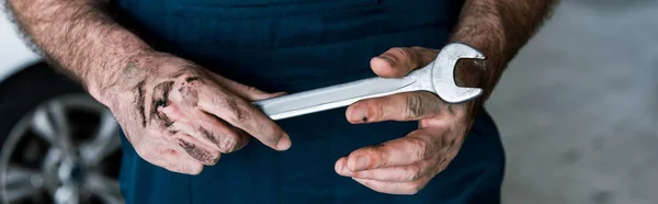 Panoramic shot  auto mechanic with mud on hands holding hand wrench in car repair station — Stock Photo