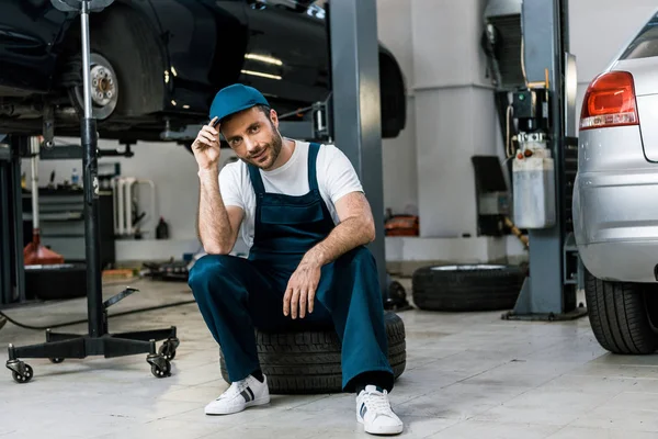 Glücklicher bärtiger Mann berührt Mütze und sitzt auf Autoreifen — Stockfoto