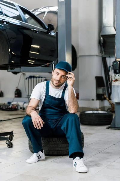 Cheerful bearded man touching cap and sitting on car tire — Stock Photo