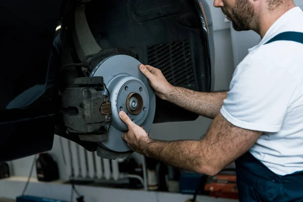 Ausgeschnittene Ansicht eines bärtigen Automechanikers, der eine metallische Autobremse in der Nähe von Automobile hält — Stockfoto