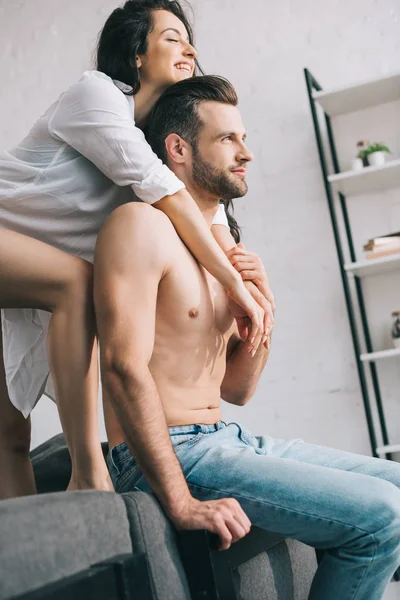 Mujer atractiva y sonriente en camisa abrazándose con hombre guapo y sin camisa - foto de stock