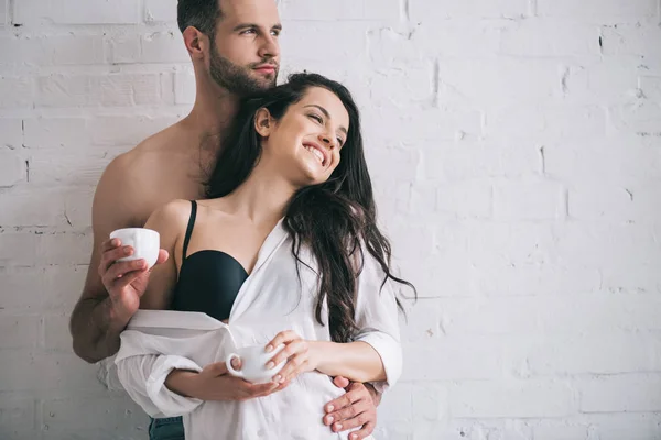 Handsome man and smiling woman in bra holding cups and looking away — Stock Photo