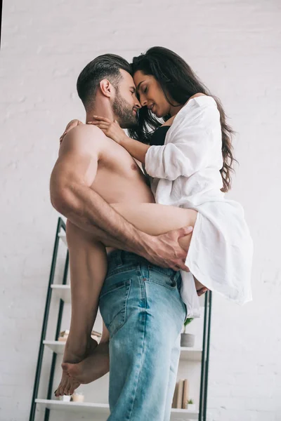 Guapo hombre en jeans sosteniendo y besando con sexy mujer en camisa - foto de stock
