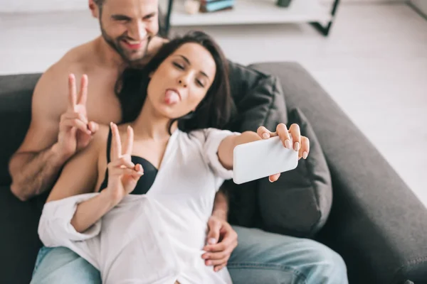 Hombre guapo y mujer sexy en camisa y sujetador mostrando signo de victoria y tomar selfie - foto de stock