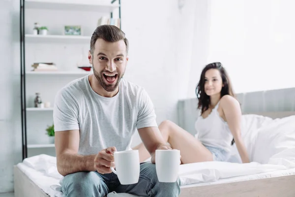 Sorrindo homem segurando copos com chá e mulher sentados na cama — Fotografia de Stock