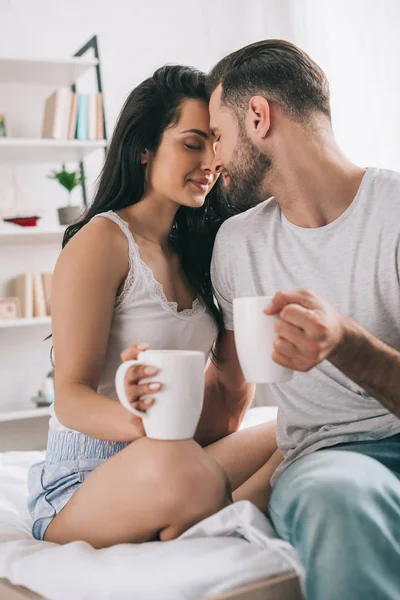 Atractiva y morena mujer con los ojos cerrados y hombre con tazas besos - foto de stock