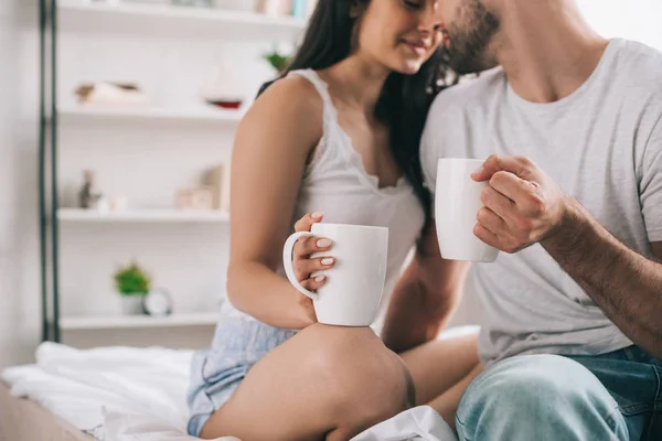 Vista recortada de hombre y mujer sosteniendo tazas con té y besos - foto de stock