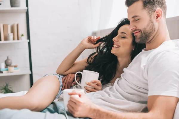 Atractiva y morena mujer y hombre sosteniendo tazas, acostado en la cama y abrazando - foto de stock