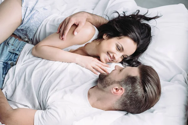 Atractiva y morena mujer y hombre sonriendo, acostado en la cama y abrazando - foto de stock