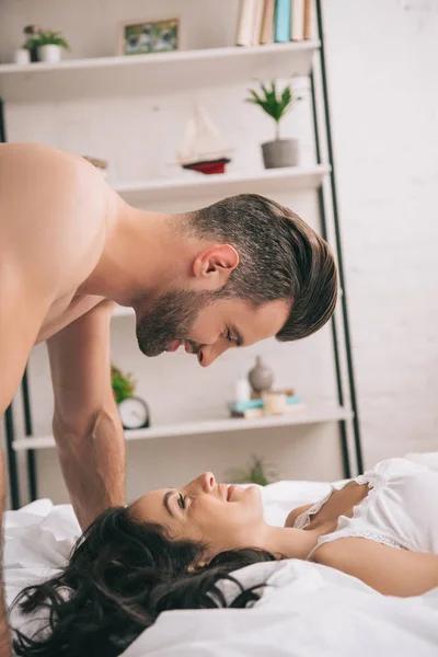 Vista lateral de homem bonito beijando mulher bonita e sorridente deitada na cama — Fotografia de Stock