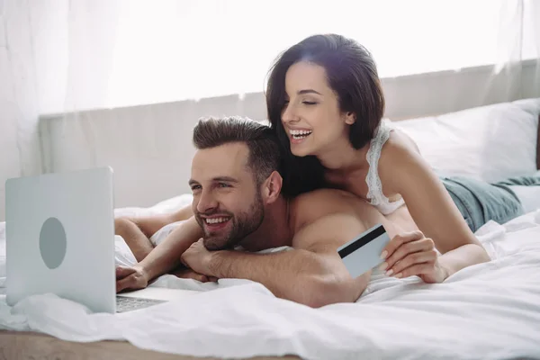 Mulher bonita e sorridente segurando cartão de crédito e homem bonito usando laptop — Fotografia de Stock