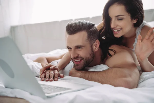 Hermosa mujer ondeando y hombre guapo usando el ordenador portátil en el dormitorio - foto de stock